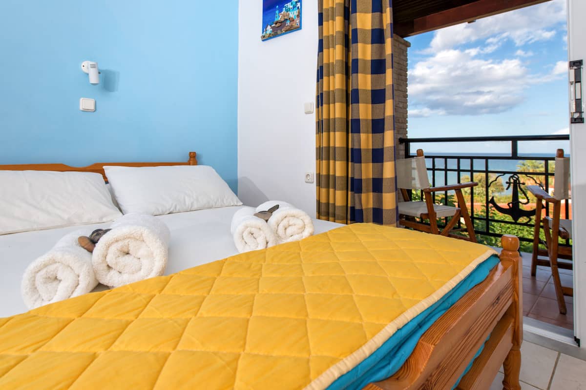 Habitación de hotel con cama, colcha amarilla acolchada, almohadas blancas y toallas enrolladas. Tiene un balcón con cortina a cuadros y vista al mar.