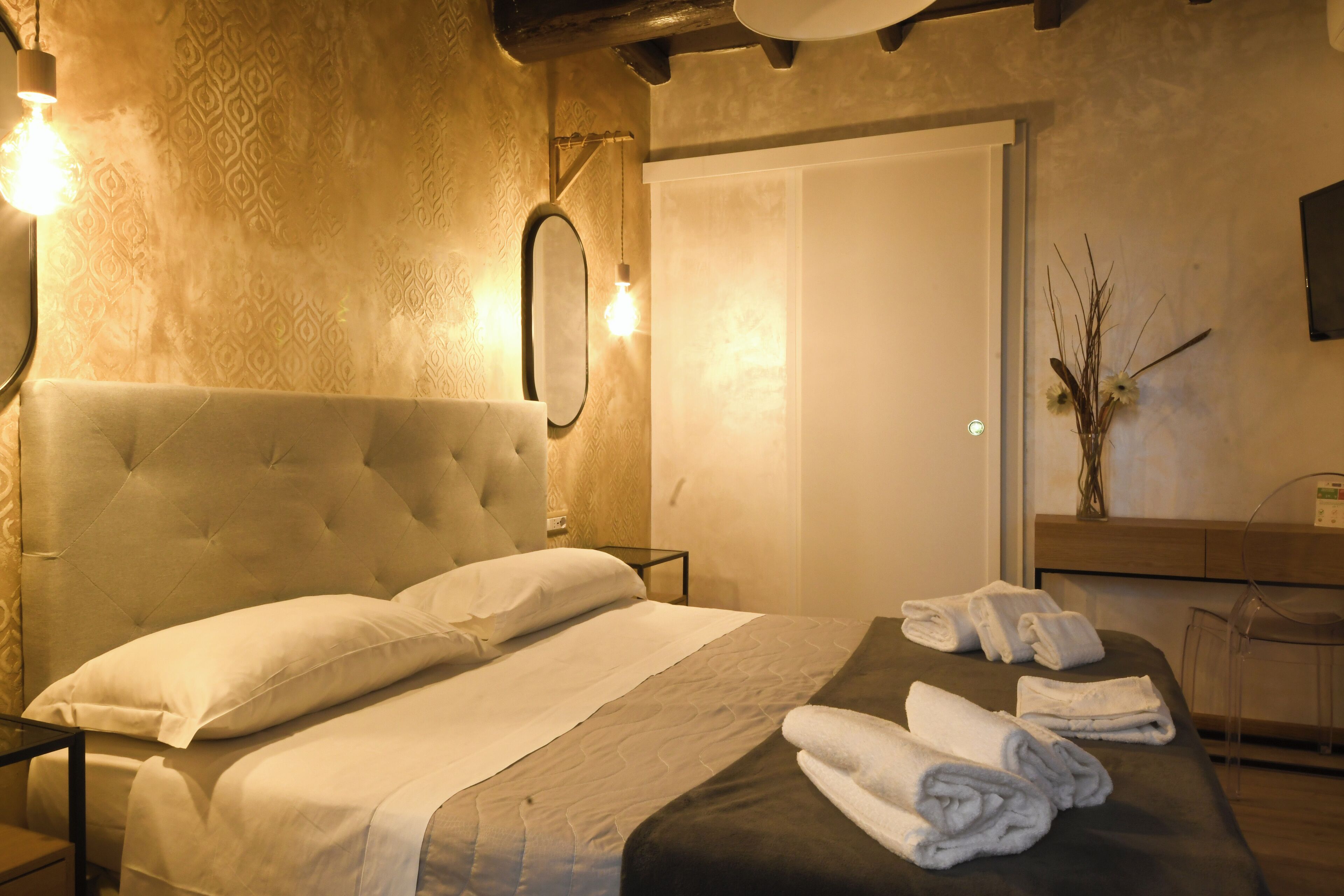 Habitación de hotel con una cama grande, una mesita de noche, un espejo, un baño con lavamanos y espejo, y un florero con flores en la pared.