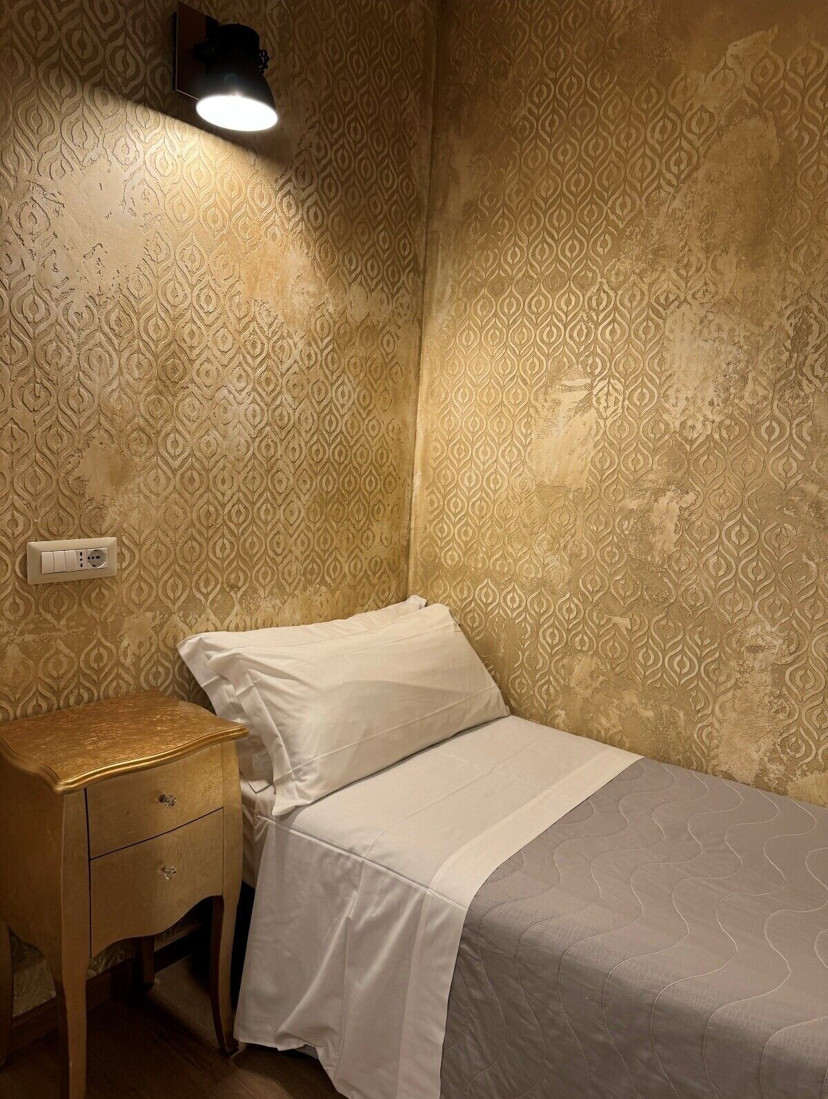 A small, dimly lit room with patterned wallpaper, a single bed, a nightstand, and a wall-mounted light.