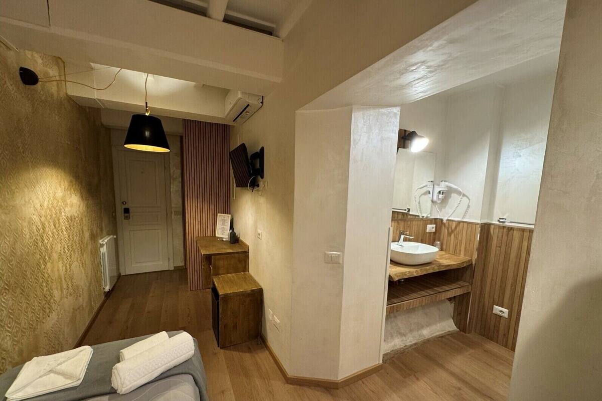 Habitación de hotel compacta con cama, una pequeña mesa de madera, un espejo y un baño con lavamanos y toallero.