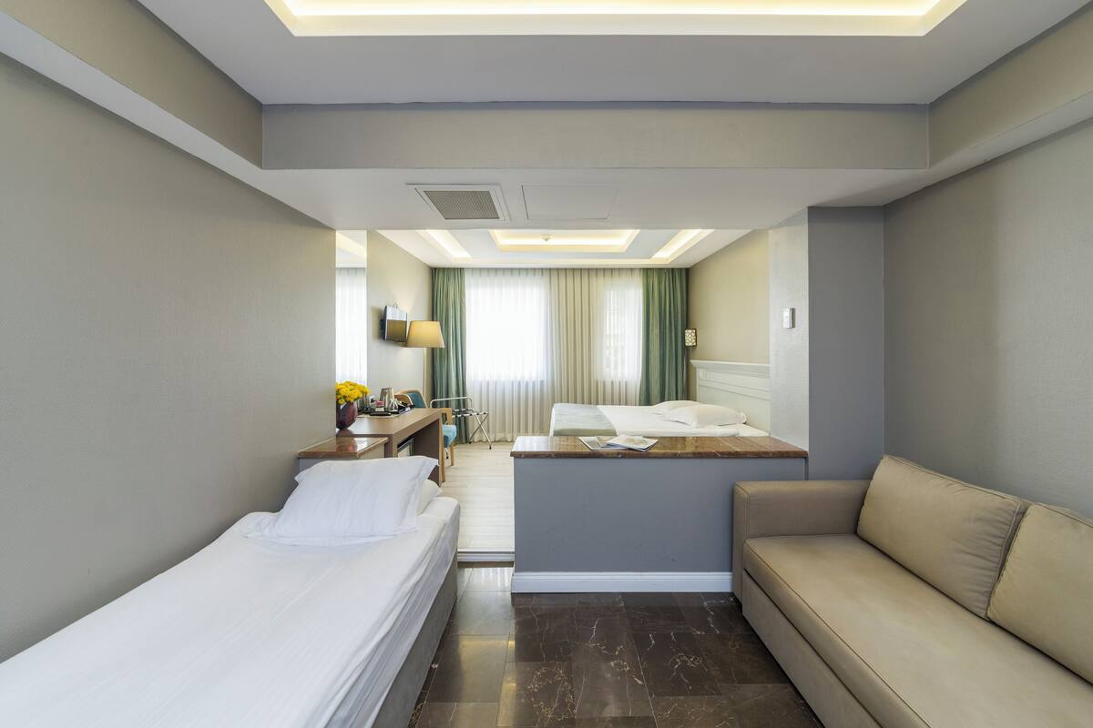 A hotel room with two beds, a sofa, a desk with a lamp, and a view of the hallway.