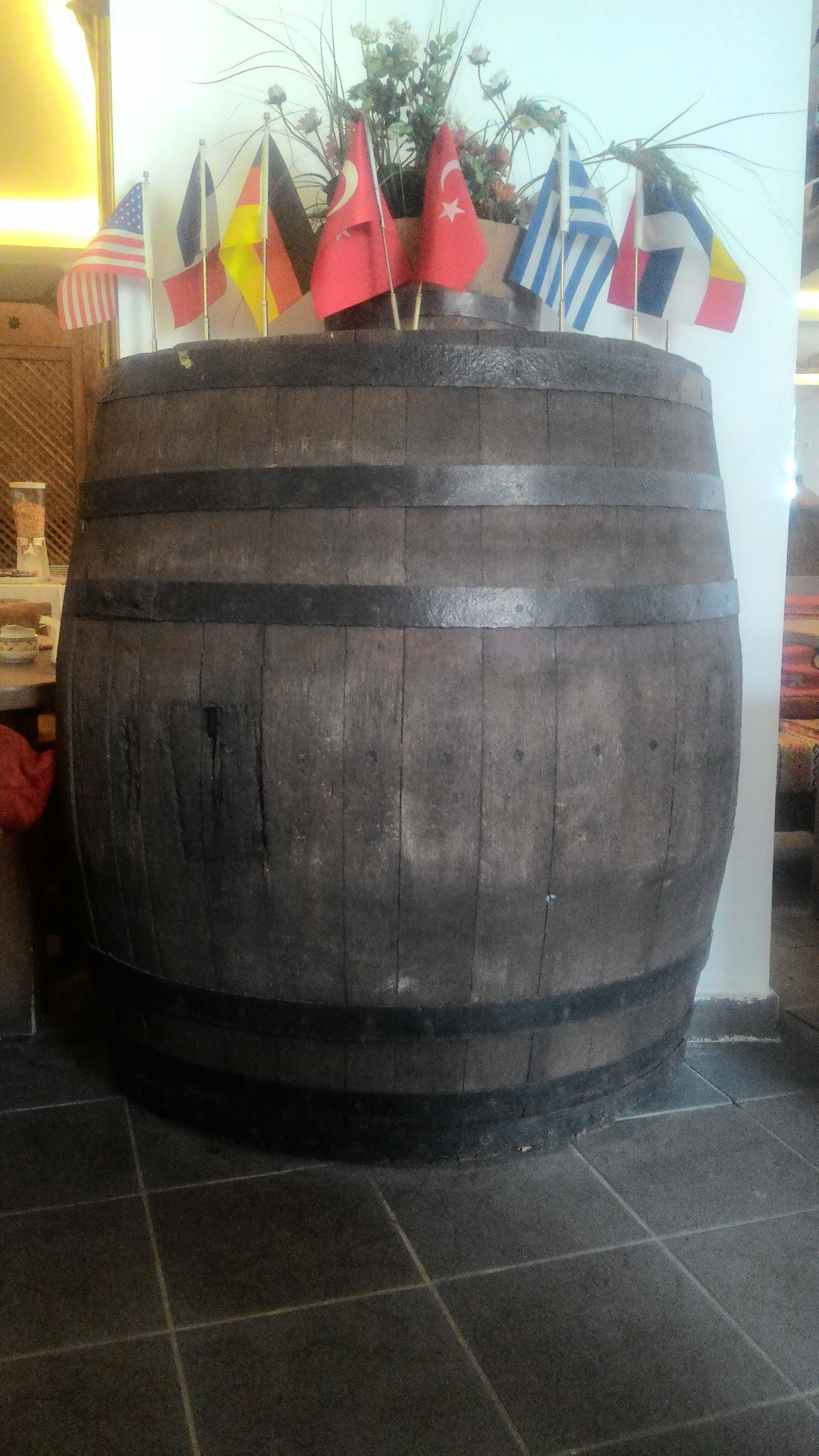 Un barril de madera grande con varias banderas en la parte superior, colocado en el interior sobre un piso de baldosas.