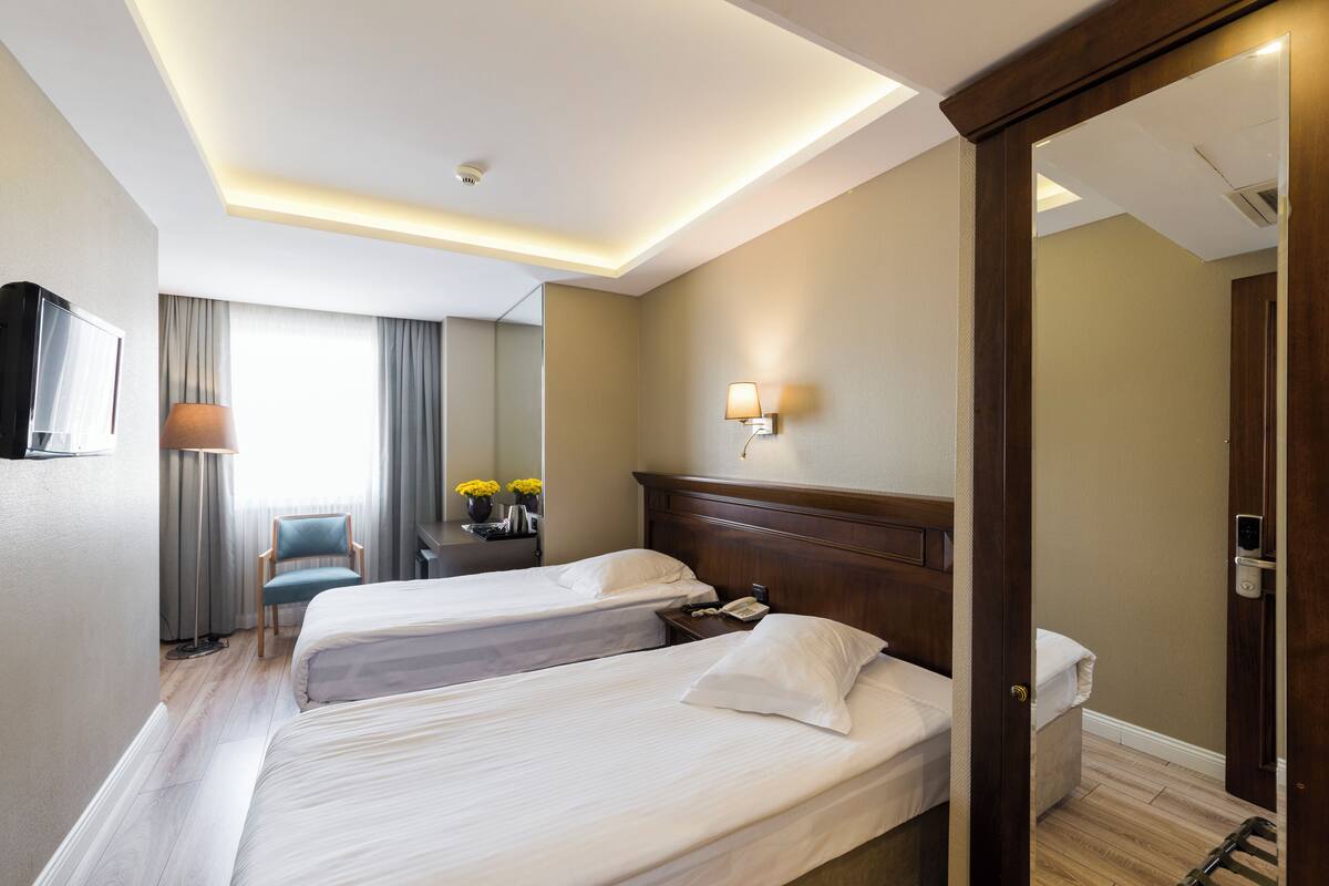 A hotel room with two beds, a desk with a lamp, a chair, and a view of the outdoors.