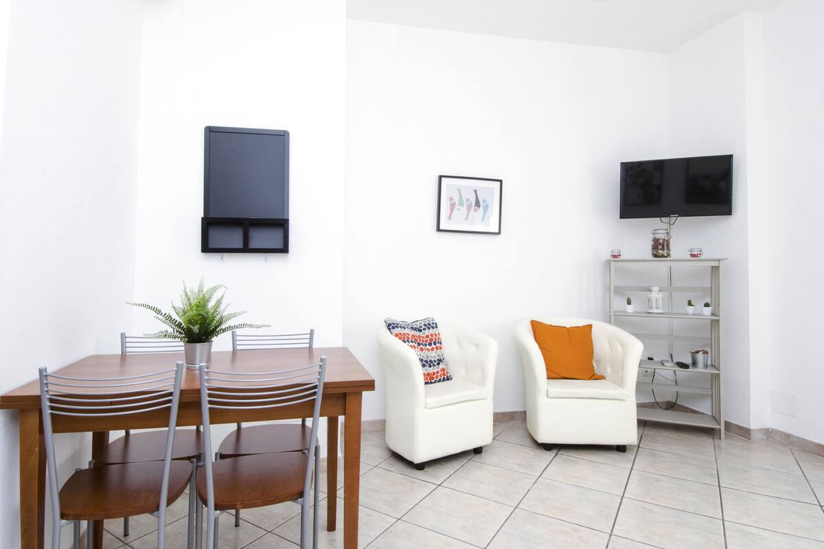 Een moderne woonkamer met een eettafel, twee stoelen, een bank, een televisie en een klein plankje met decoratieve objecten.