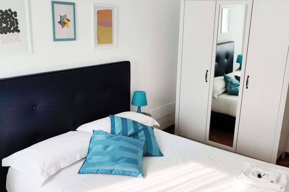 Een opgemaakt bed met blauwe kussens, een witte kledingkast en een kleine tafel met een lamp.