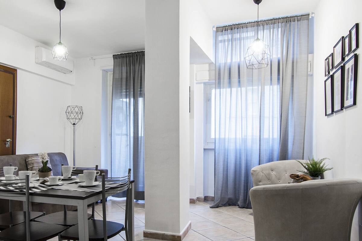 Een moderne woonkamer met een eethoek, met een bank, een eettafel met stoelen en een groot raam met gordijnen.
