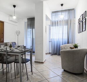 A modern living room with a dining area, featuring a sofa, a dining table with chairs, and a large window with curtains. - OM2Rome (Rome)