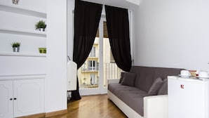 A compact living space with a sofa, a small kitchenette, a window with curtains, and a balcony view. - OM2Rome (Rome)