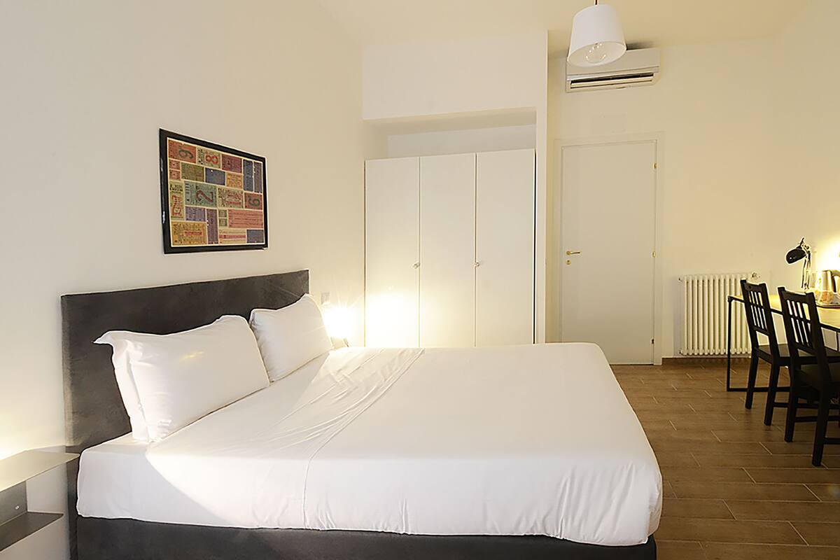 Een net opgemaakt bed met witte lakens, een ingelijst schilderij aan de muur, een kledingkast en een eethoek met stoelen.