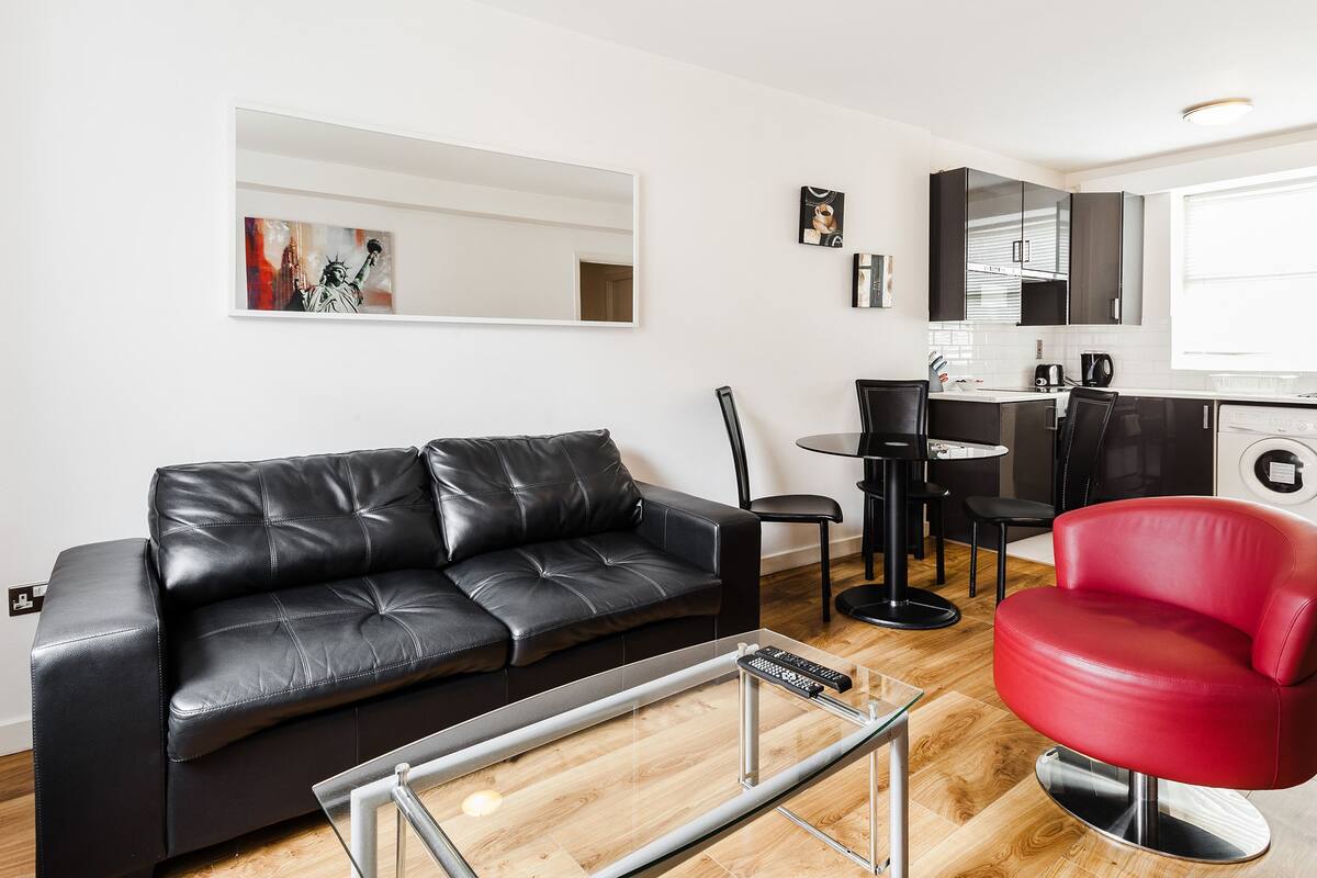 A modern living room with a black leather sofa, a red chair, a glass coffee table, and a small dining area with a black table and chairs. A washing machine is visible in the background.