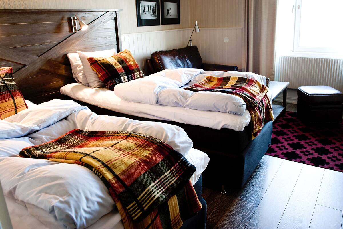 Een hotelkamer met twee bedden, een houten hoofdeinde, een zwarte leren fauteuil, een klein tafeltje en een raam met gordijnen.