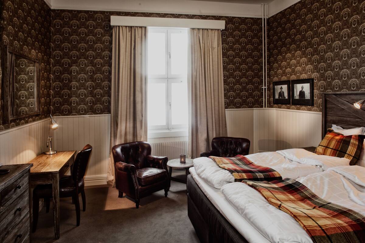 Een hotelkamer met een houten bureau, twee leren fauteuils, een klein rond tafeltje, een bed met een ruitdeken en een raam met gordijnen.