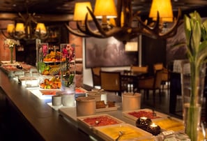 Ein reichhaltiges Buffet mit verschiedenen Speisen und Getränken, darunter Früchte, Desserts und Erfrischungen, auf einer langen Tafel mit angenehmer Beleuchtung.