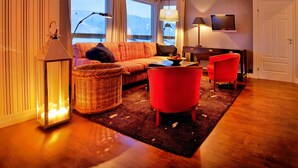Ein gemütliches Wohnzimmer mit einer Couch, zwei roten Sesseln, einem Tisch, einem Fernseher und einem großen Fenster mit Blick.