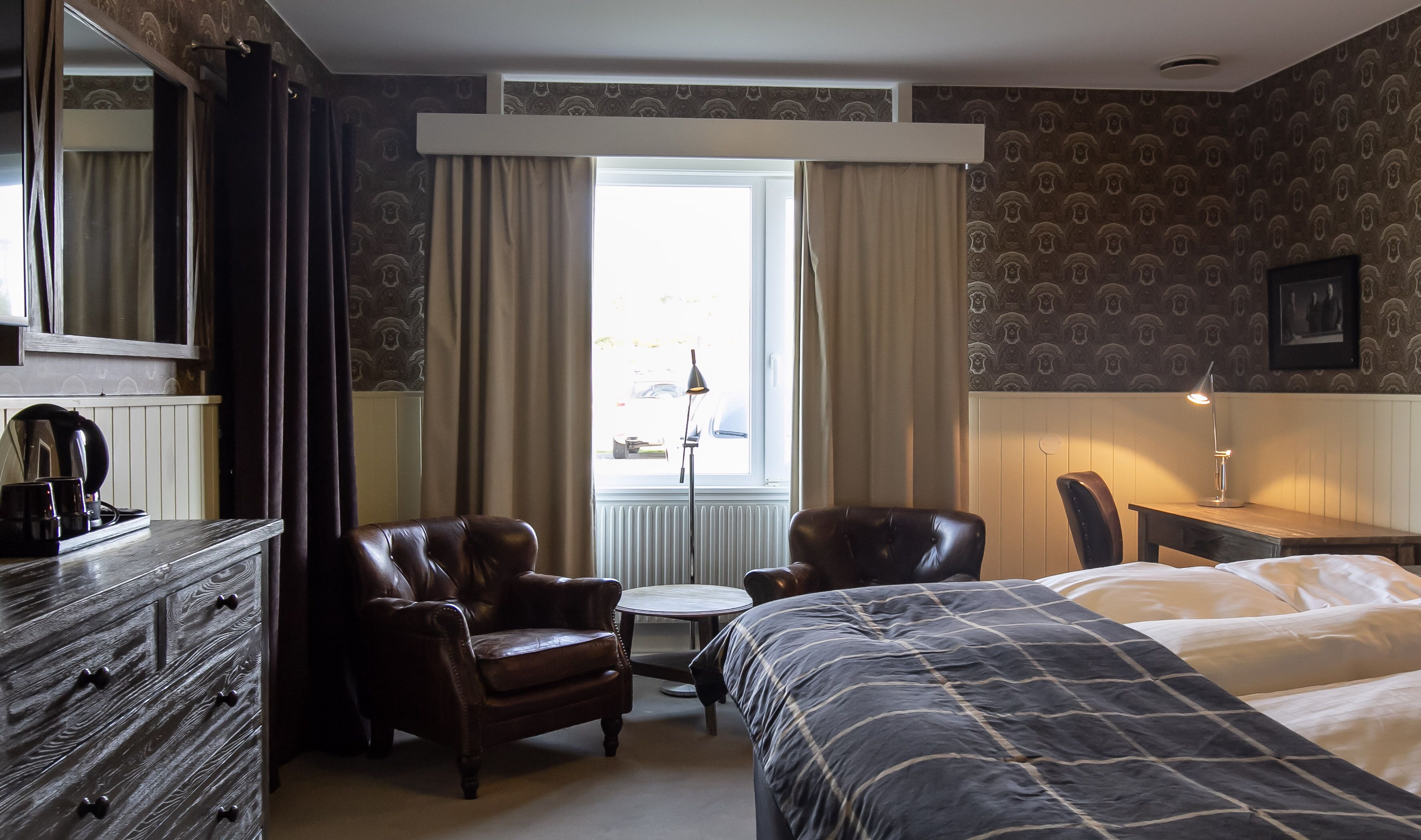 Et hotelværelse med seng, to stole, et lille bord, en kommode og et vindue med gardiner.