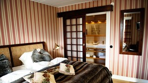 A bedroom with striped wallpaper, a wooden headboard, a bed with a dark blanket and decorative pillows, a bedside lamp, and a mirror on the wall.
