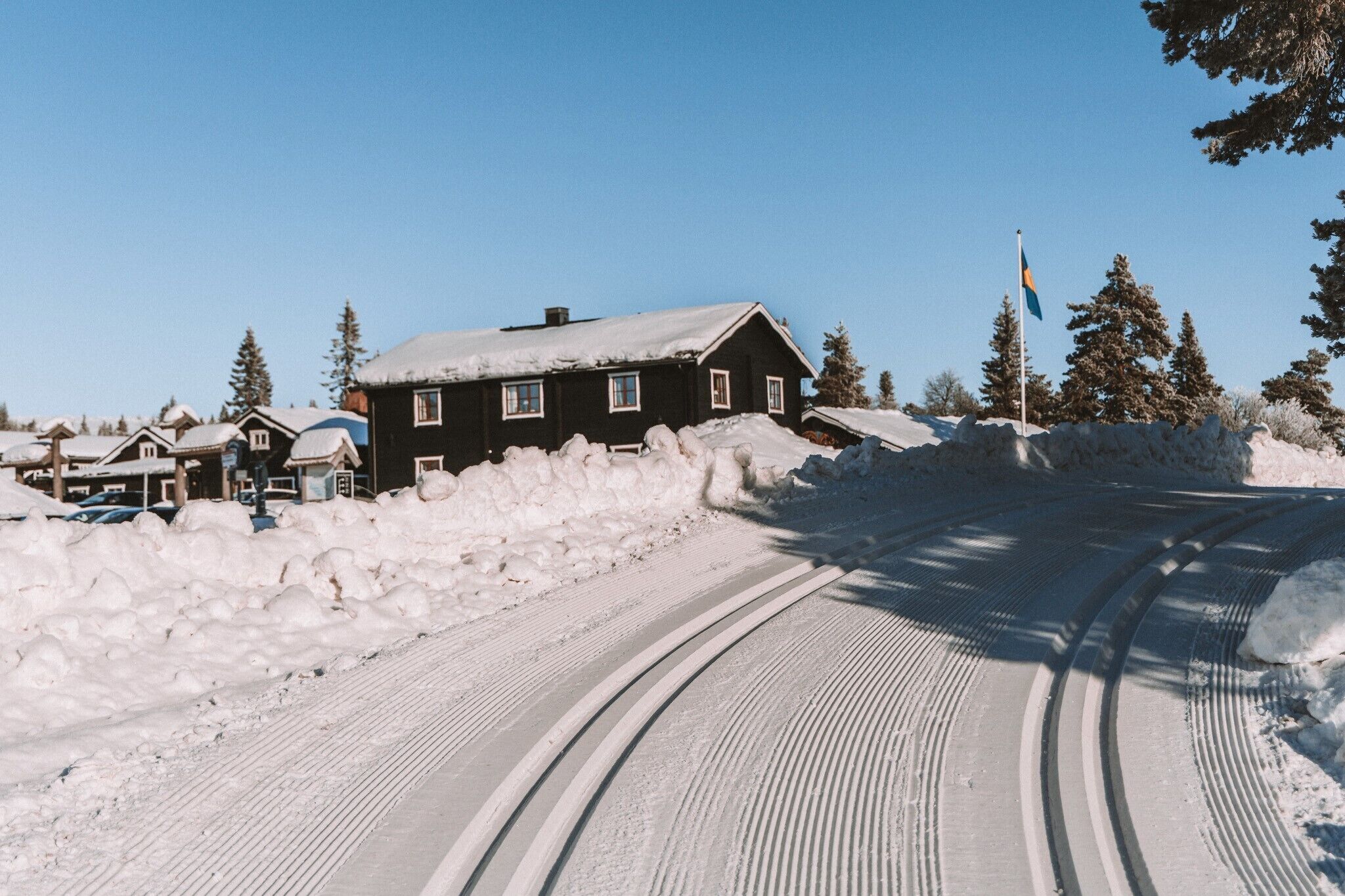 A snow-covered road with tire tracks leading to a black house with a flag.
