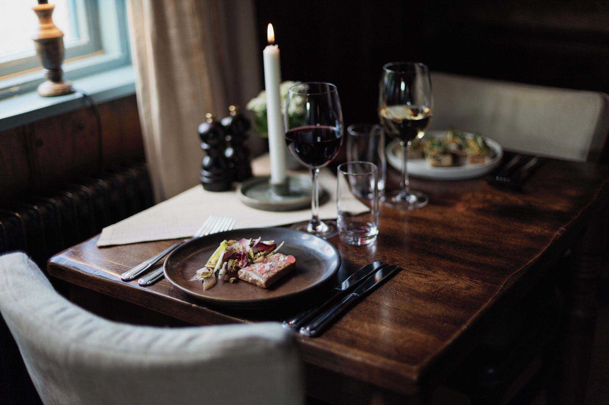 A dining table set with plates, glasses, and cutlery, a candle, and a window with curtains.