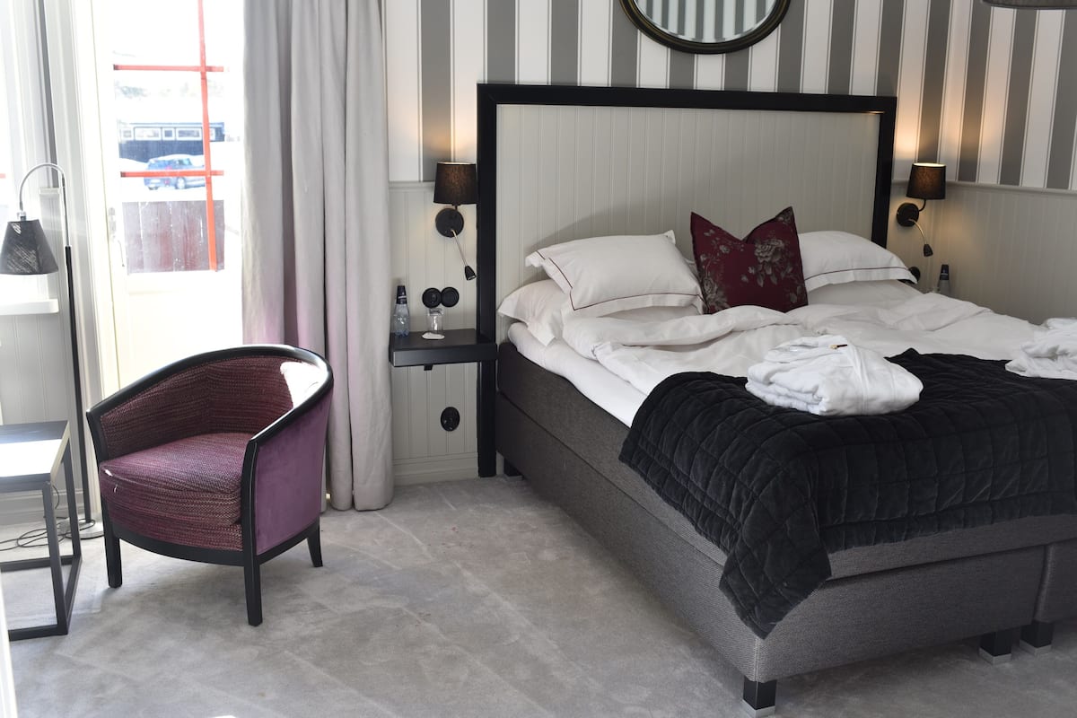 Un lit bien fait, avec une tête de lit rayée en noir et blanc, un oreiller rouge et un plaid noir. On y voit une chaise avec un coussin violet, une petite table avec une lampe et une fenêtre avec des rideaux.