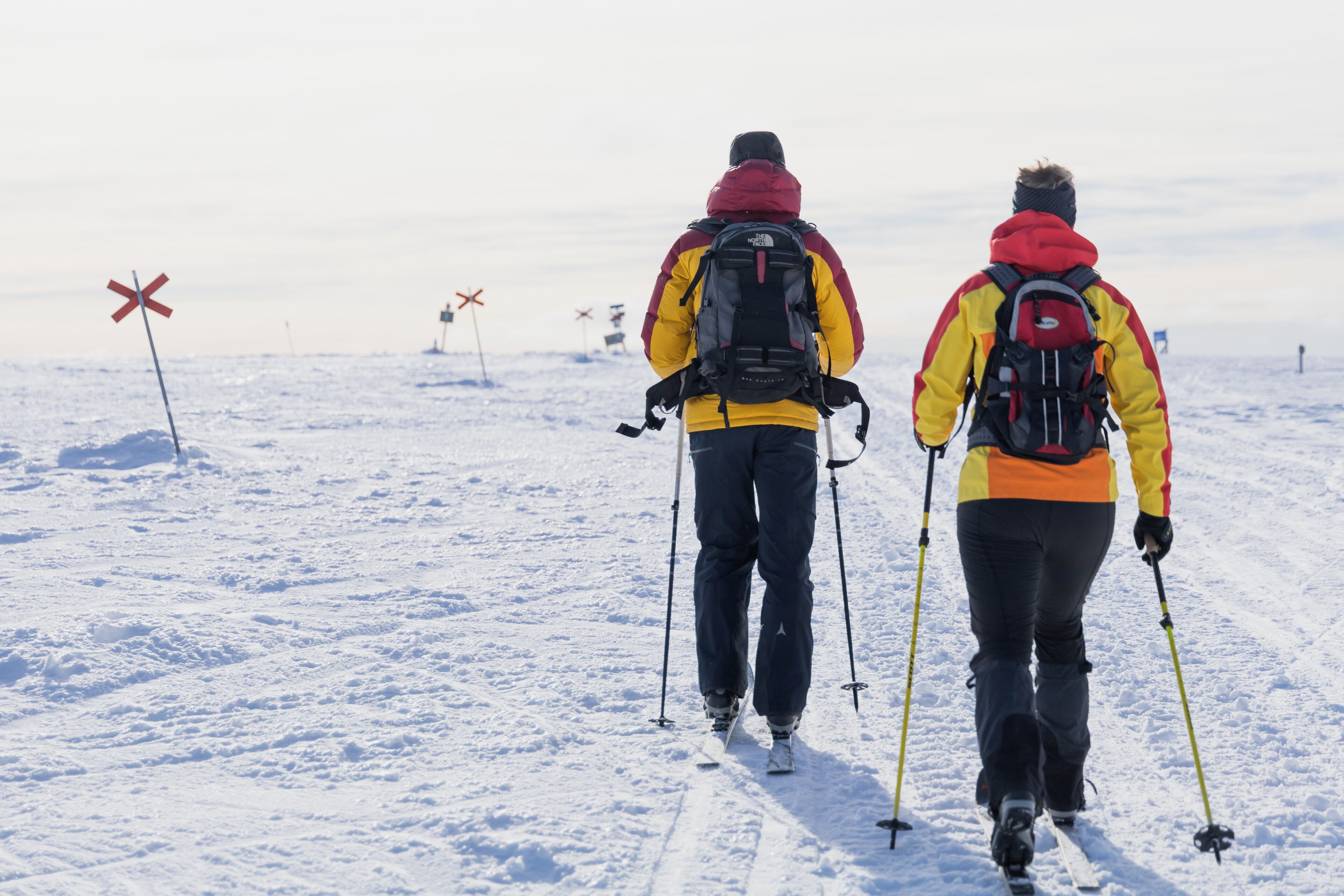 Two people walking on a snowy path with ski poles, wearing backpacks and winter clothing.