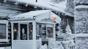 A snow-covered wooden cabin with a porch, a small covered seating area, and a snow-covered tree.