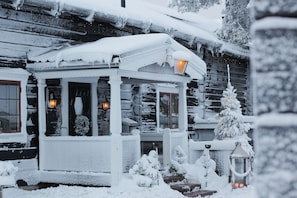 Ein mit Schnee bedecktes Holzhäuschen mit Veranda, einer kleinen überdachten Sitzgelegenheit und einem schneebedeckten Baum.