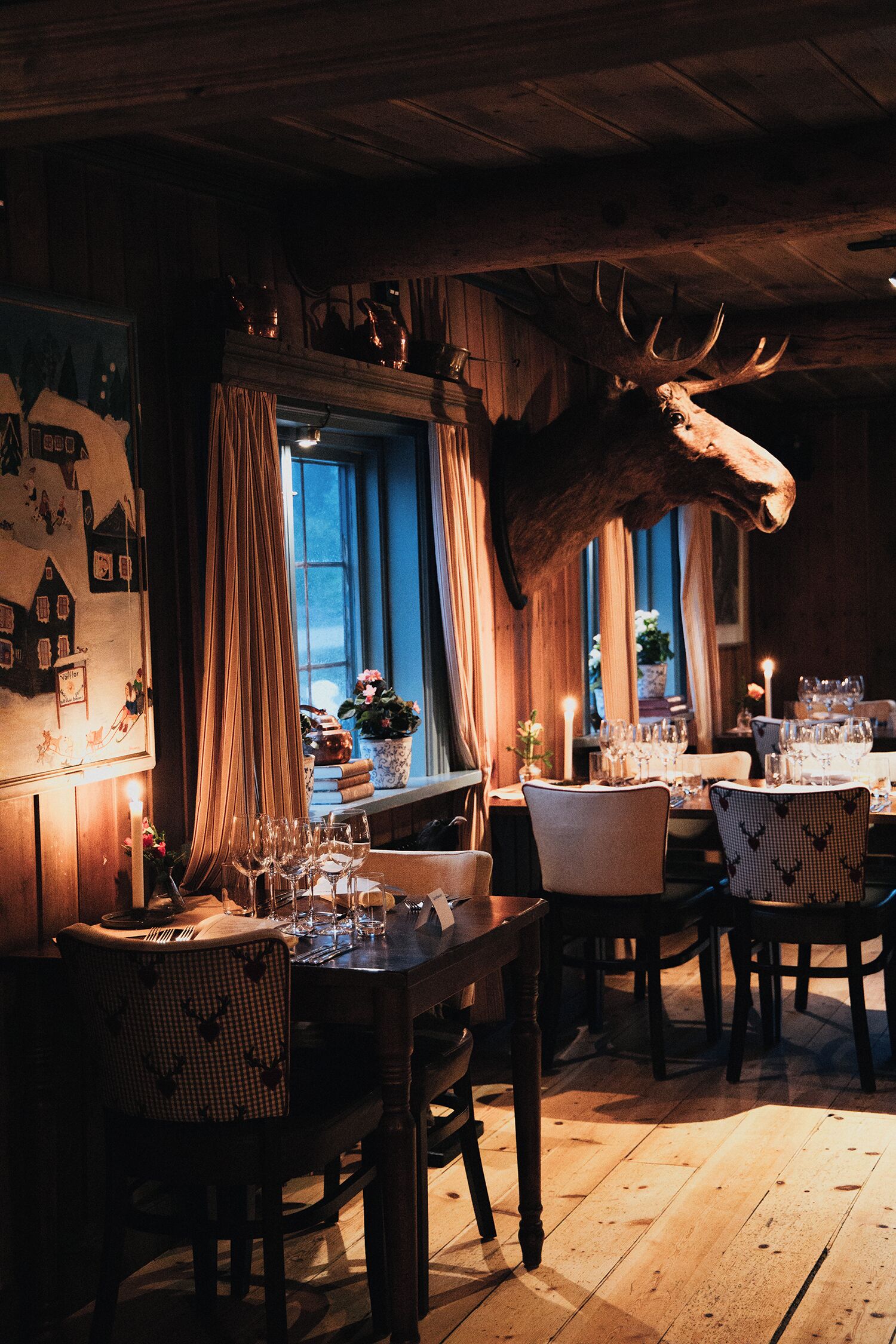 A dining area with wooden tables and chairs, a large antler decoration, and a window with curtains.