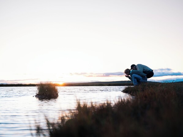 A sunset over a calm body of water with two people crouching near the edge. - Gammelgården Hotell (Salen)