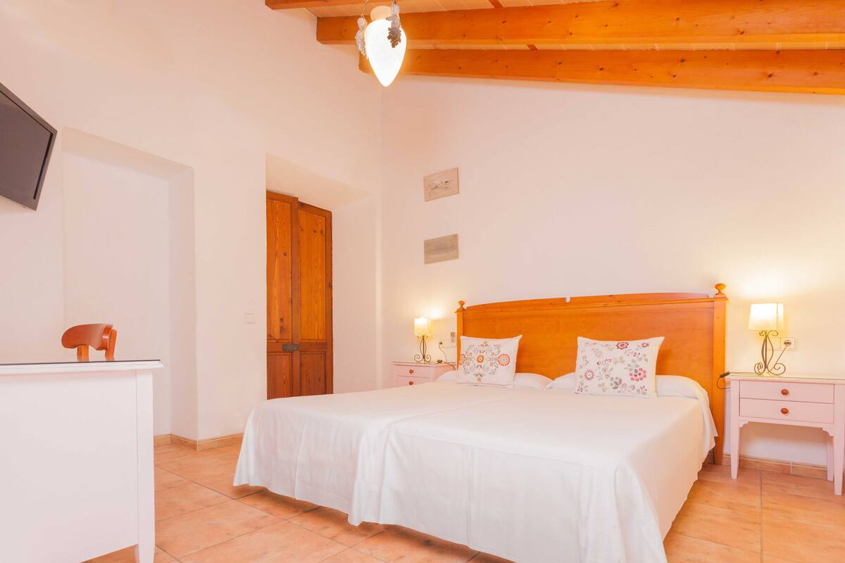 Un dormitorio con cabecera de madera, ropa de cama blanca, dos mesitas de noche con lámparas y un televisor en la pared.
