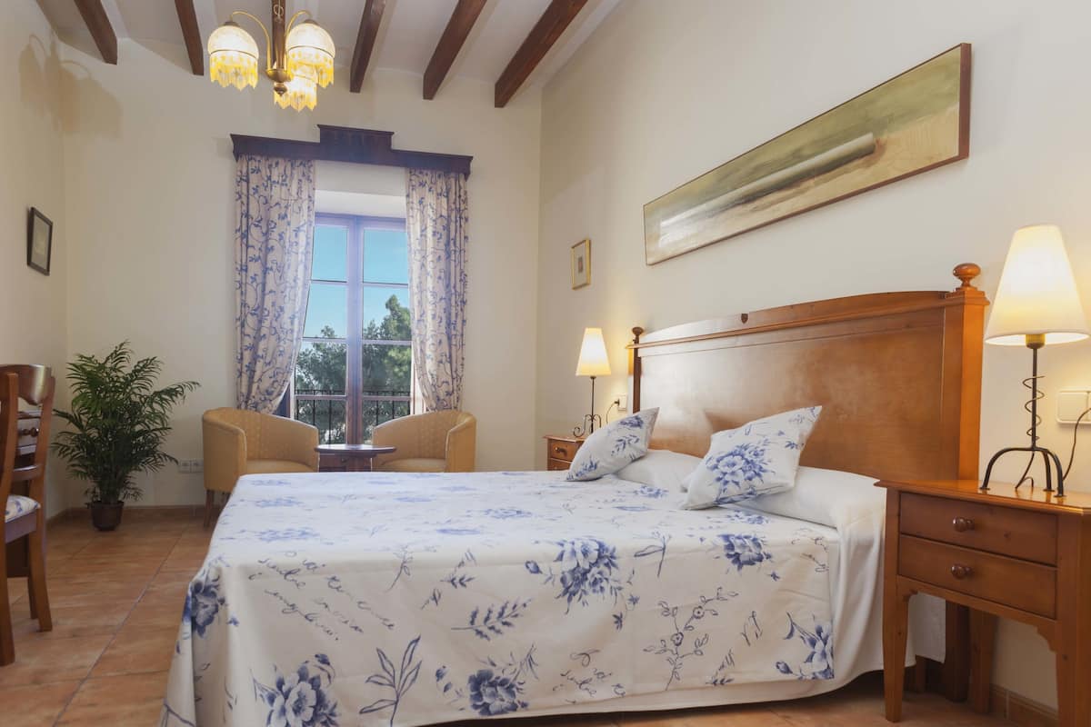 Un dormitorio con una cama grande, dos mesitas de noche con lámparas, un cabecero de madera, una ventana con cortinas, una planta en maceta y una silla.