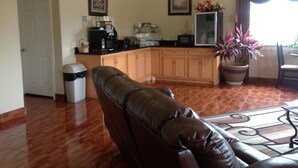 A living room with a brown leather sofa, a kitchen area with wooden cabinets, a coffee machine, and a potted plant. - Scottish Inn & Suites (Houston)