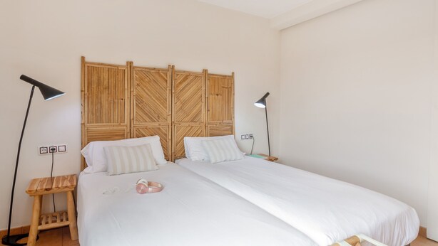 A bedroom with a wooden headboard, two bedside tables with lamps, a bed with white bedding, and a small wooden stool.