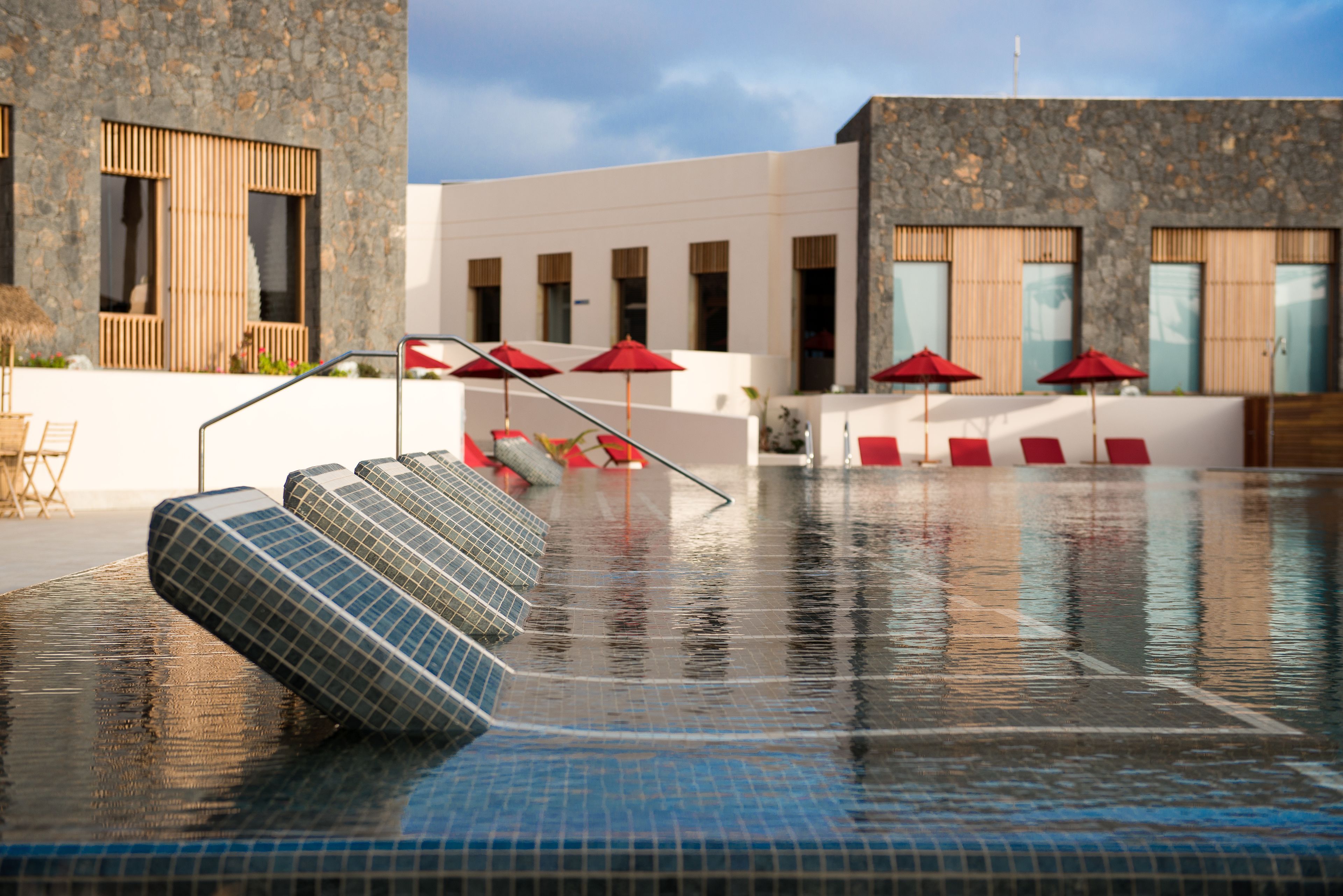 Una piscina con escalones de acceso, rodeada de sillas de descanso y sombrillas rojas.