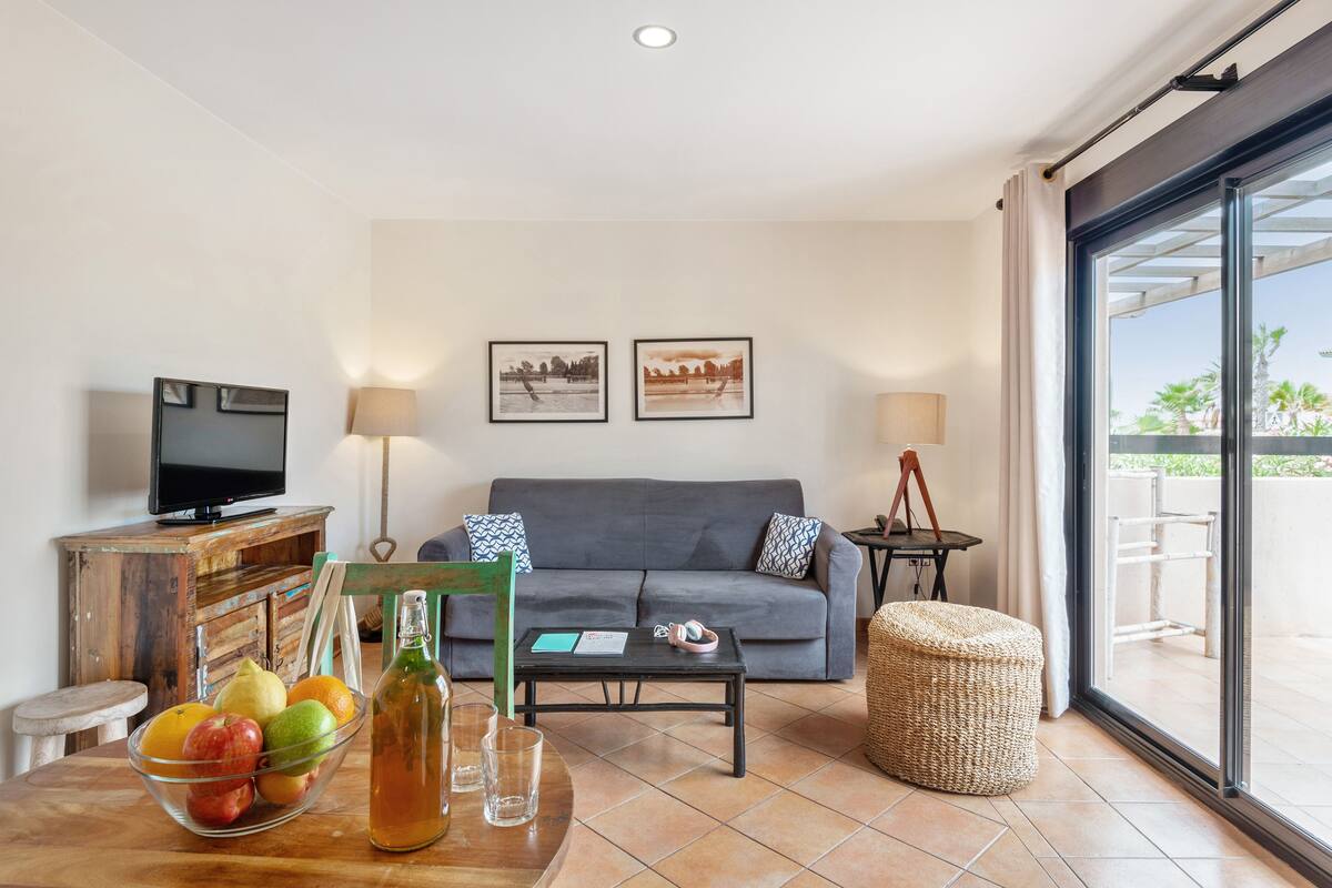 Un salon comprenant un canapé, un téléviseur, une table basse avec un saladier de fruits et un pouf en osier.