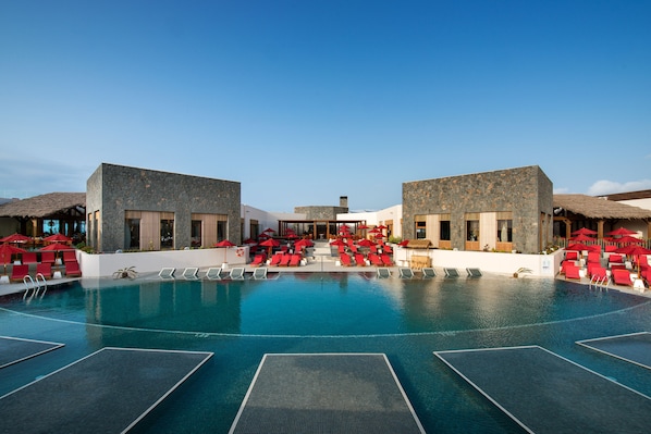 Zona de piscina con sillas reclinables rojas y sombrillas, rodeada de edificios de piedra con amplias ventanas.