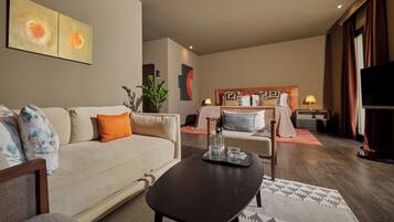 A hotel room with a sofa, a coffee table, a bed with a headboard, a TV, and a plant.