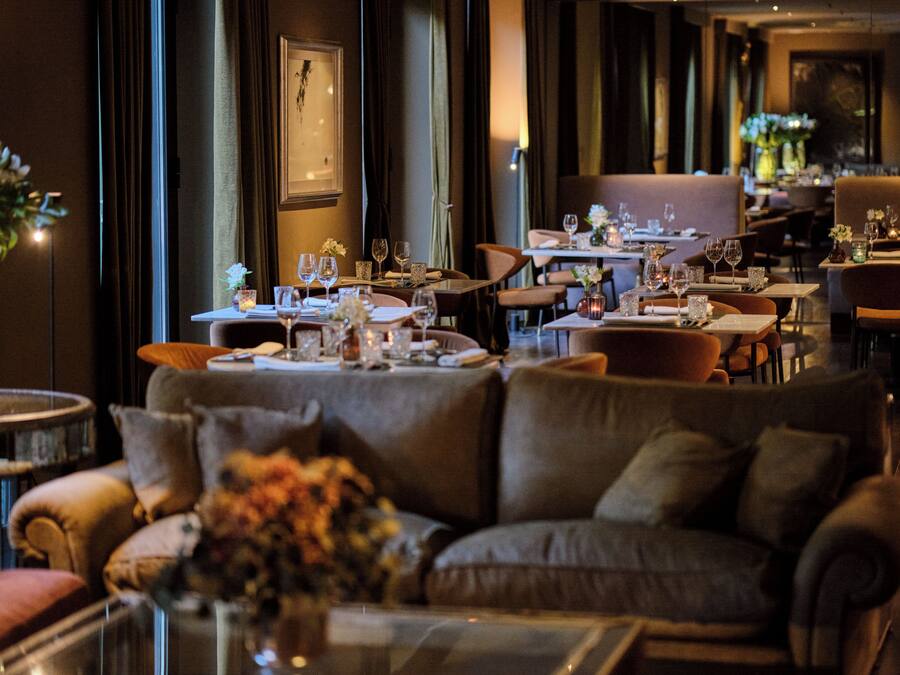 A dimly lit restaurant with tables set for dining, a sofa in the foreground, and a large window with curtains.