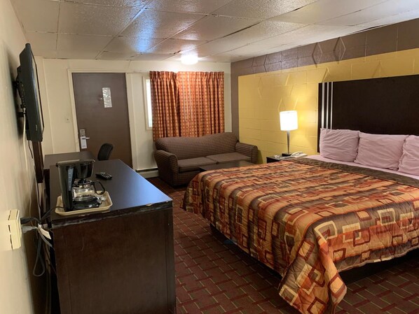 Habitación de hotel con cama, escritorio con cafetera, silla, televisor y lámpara.