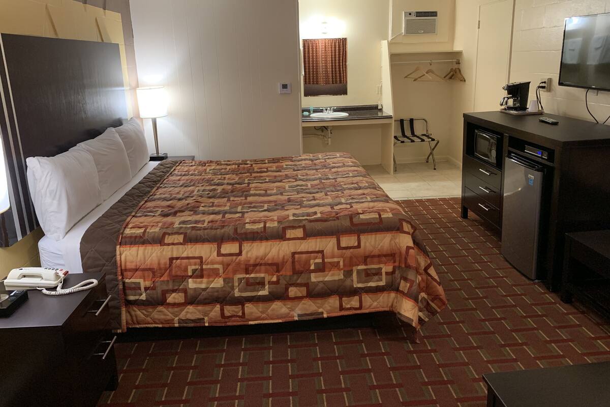 A hotel room with a large bed, a desk with a phone, a refrigerator, a TV, and a small kitchenette area.