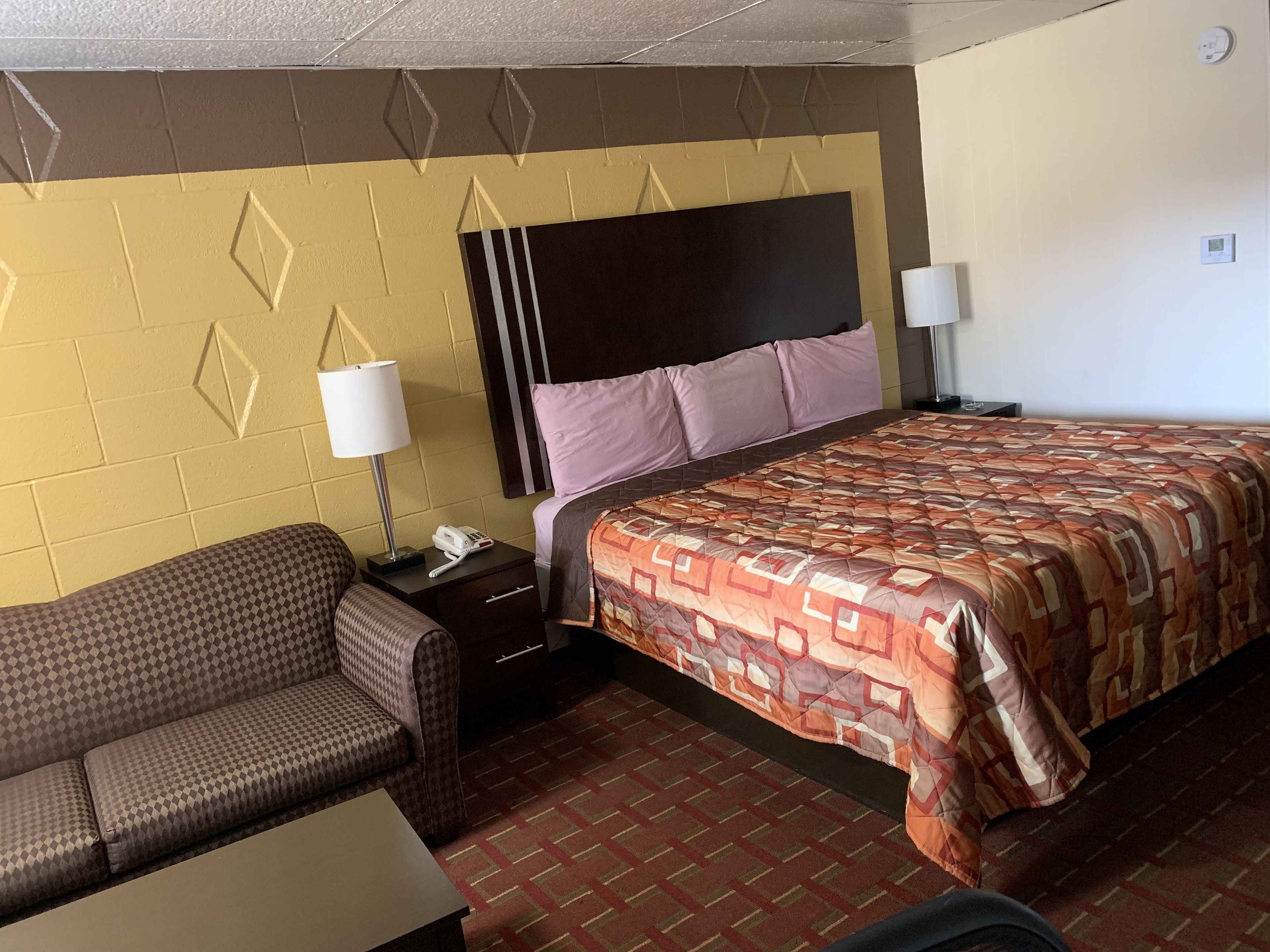 A hotel room with a bed, a sofa, a nightstand with a lamp, and a wall clock.