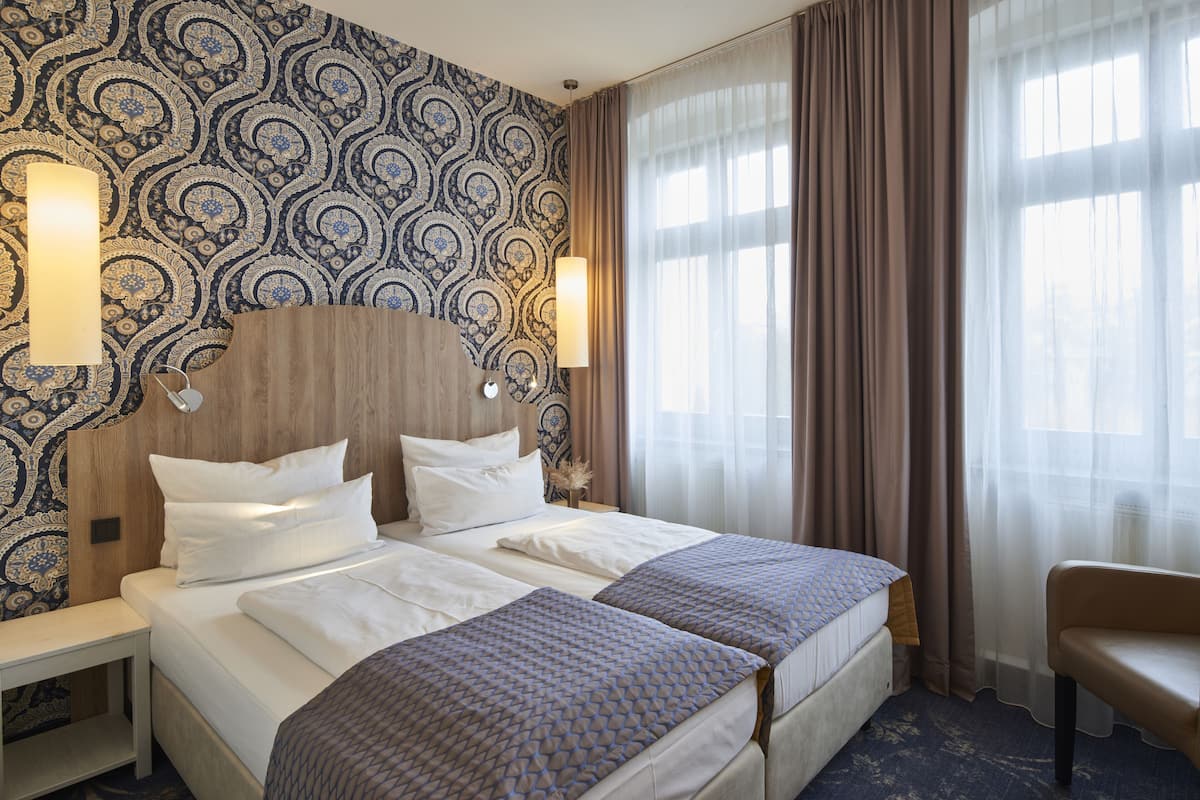 A hotel room with two beds, a nightstand, a lamp, and a chair.
