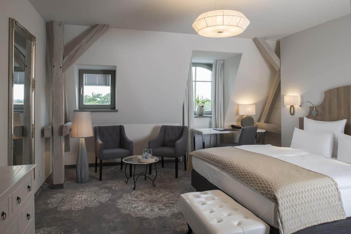 A hotel room with a bed, two armchairs, a small table, a lamp, and a window with curtains.