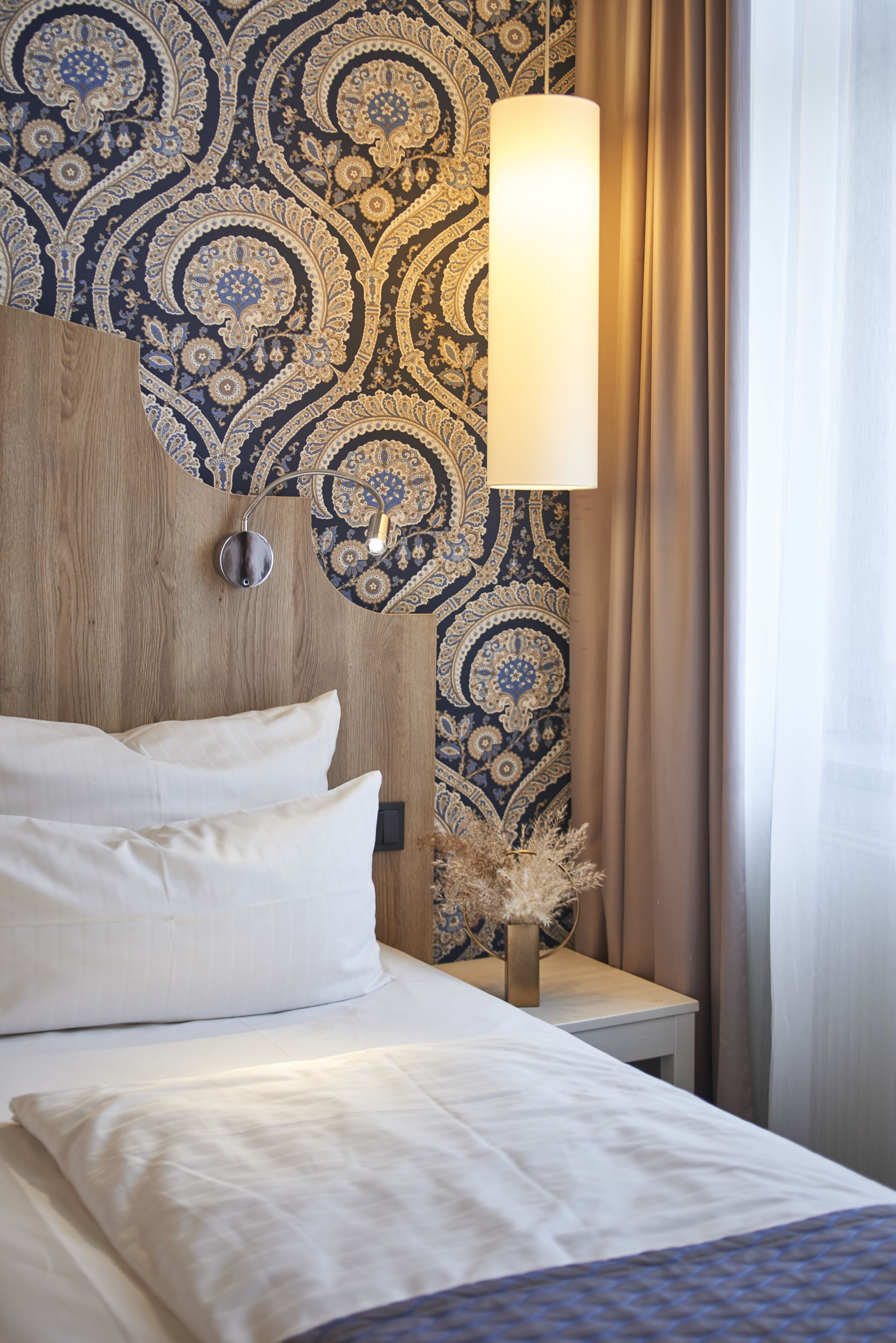 Een net opgemaakt bed met witte lakens en een houten hoofdbord, een wandlamp en behang met een patroon.