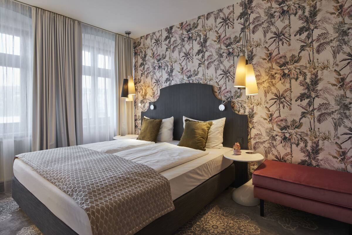 A double bed with a patterned bedspread, a headboard with two wall-mounted lights, a small bedside table with a lamp, and a red bench.