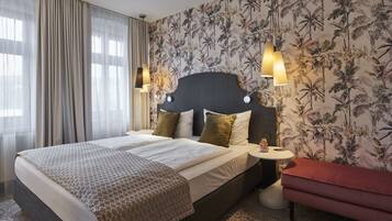 A double bed with a patterned bedspread, a headboard with two wall-mounted lights, a small bedside table with a lamp, and a red bench.