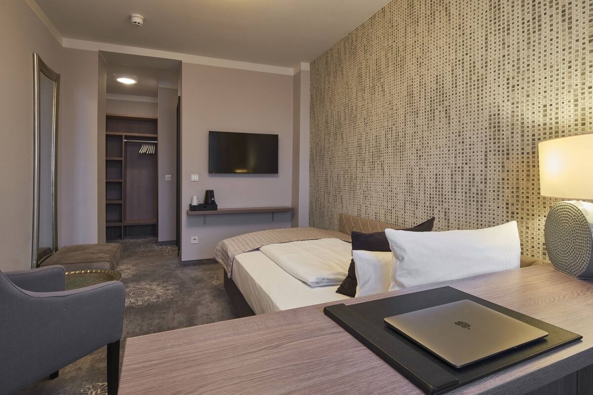 A hotel room with a bed, a desk with a laptop, a TV on the wall, and a patterned wall.