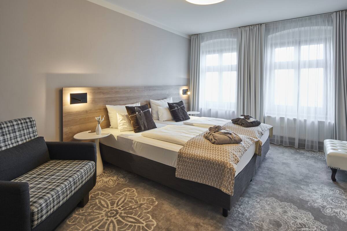 A modern hotel room with a large bed, a sofa, a small table, and a large window with sheer curtains.