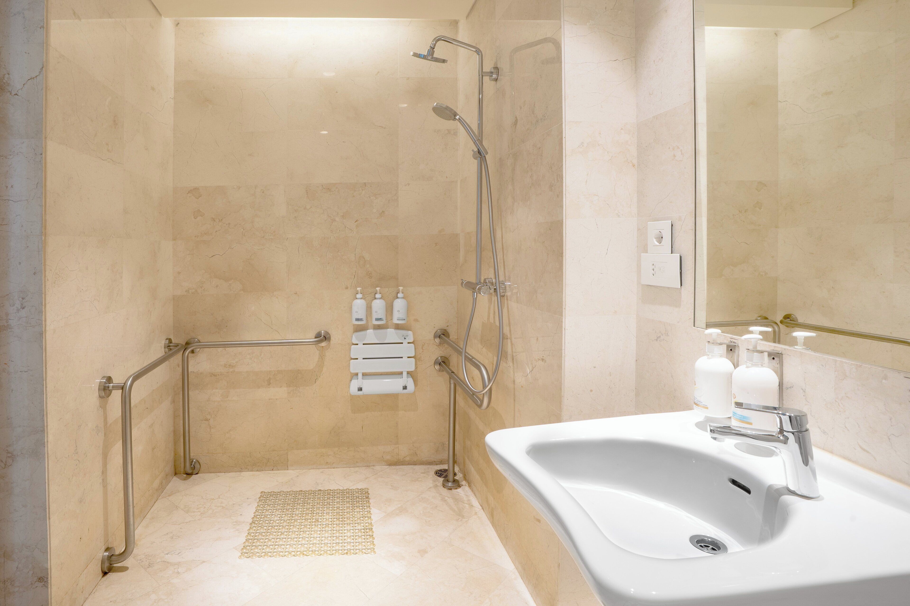 A modern bathroom with a walk-in shower, grab bars, a sink with a mirror, and hand soap dispensers.
