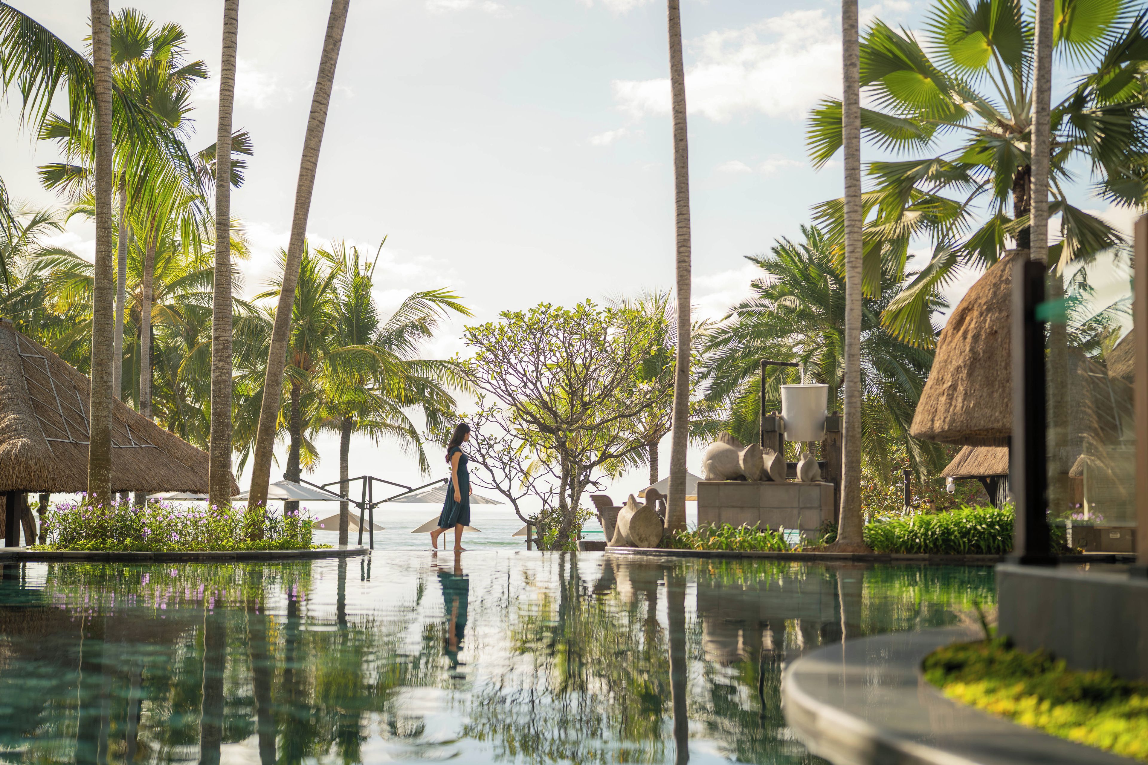 A resort pool area with palm trees, a thatched-roof structure, and a clear view of the ocean.
