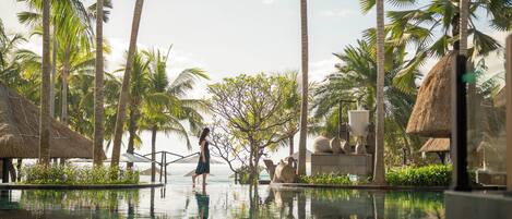 A resort pool area with palm trees, a thatched-roof structure, and a clear view of the ocean.